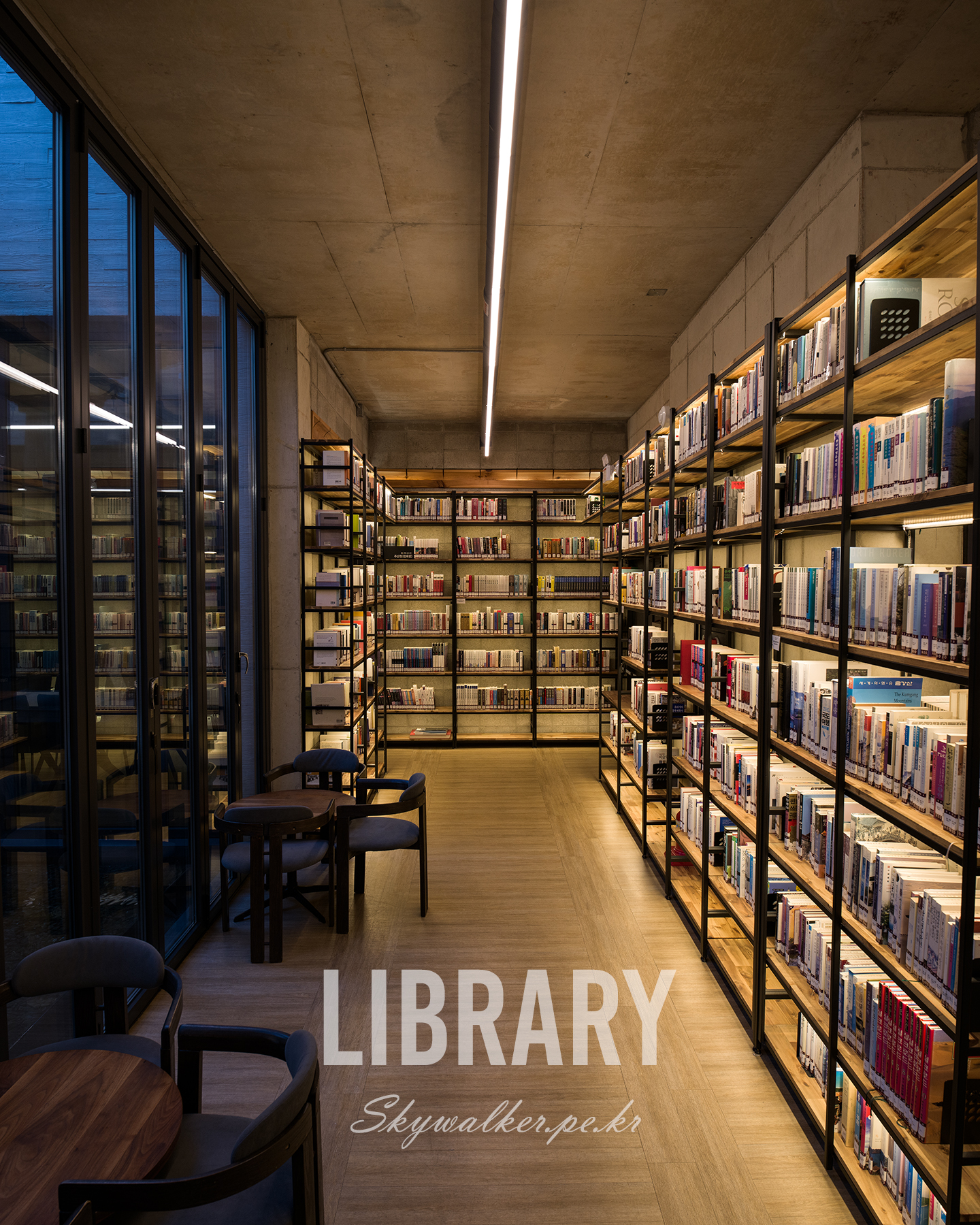 Library - A quiet library aisle with tall bookshelves, glass walls, warm shelf lighting, and a linear ceiling light creating a modern interior mood. - #Library #Bookshelves #InteriorPhotography #ModernArchitecture #WarmLight #ReadingRoom #Bookstacks #ConcreteCeiling #QuietSpace #Symmetry
