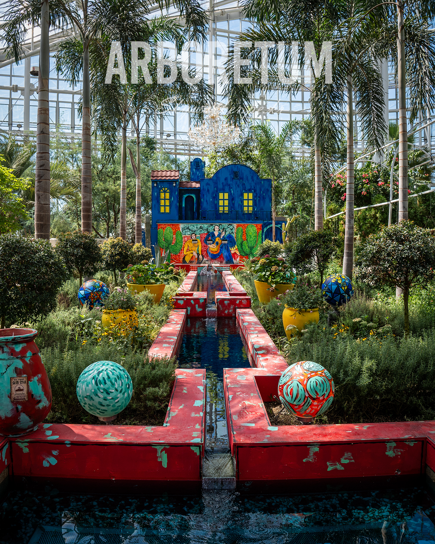 Arboretum - Sunlit greenhouse garden with palms frames a colorful water feature and painted blue facade under a glass roof. - #arboretum #greenhouse #palmtrees #botanicalgarden #waterfeature #gardenart #glassroof #colorful #indoorGarden #tropicalPlants