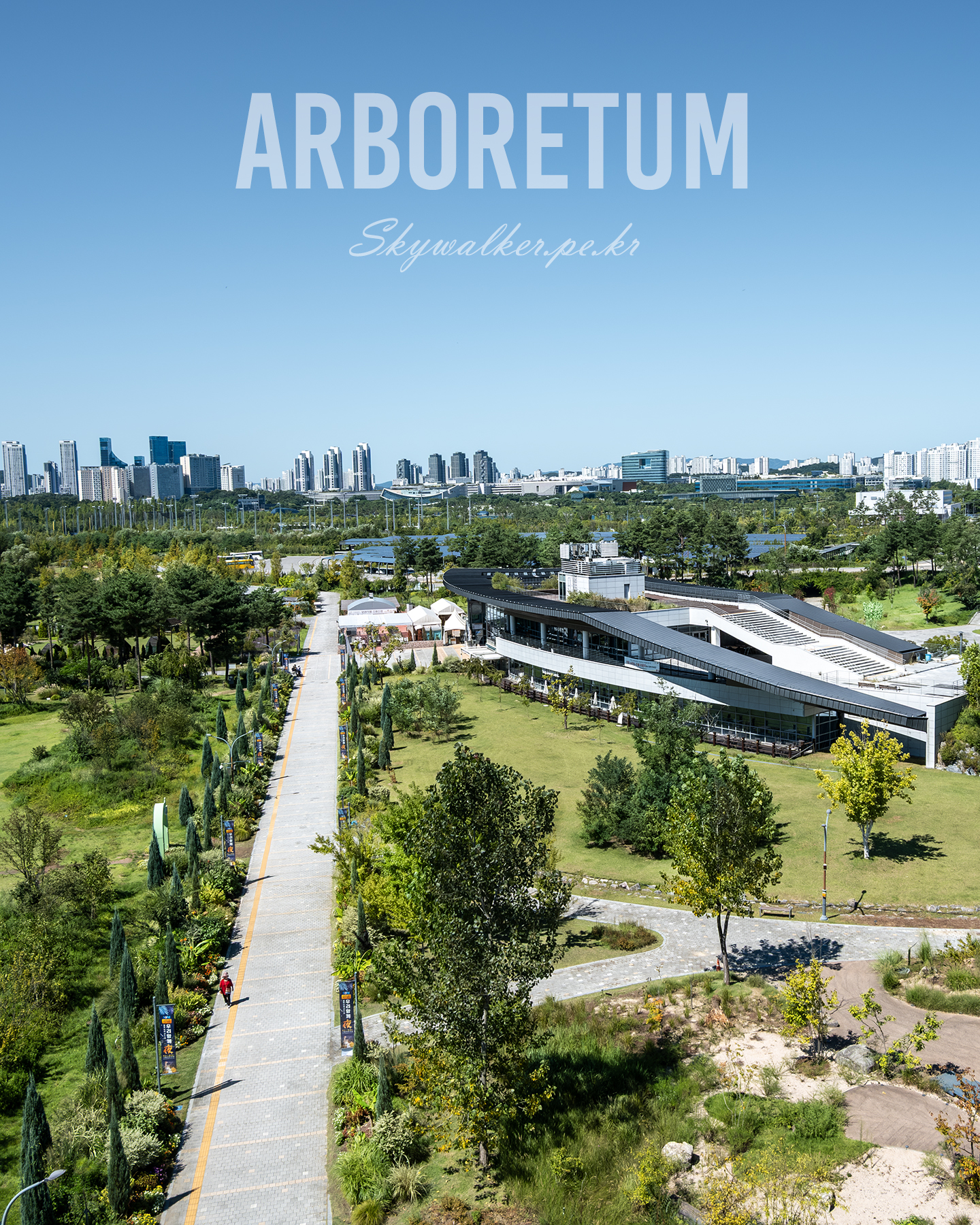 Arboretum - An elevated view shows a green arboretum with walking paths, a modern building, and a distant city skyline under clear blue sky. - #Arboretum #UrbanNature #CitySkyline #GreenSpace #GardenPath #ClearSky #LandscapePhotography #ModernArchitecture #AerialView #SunnyDay