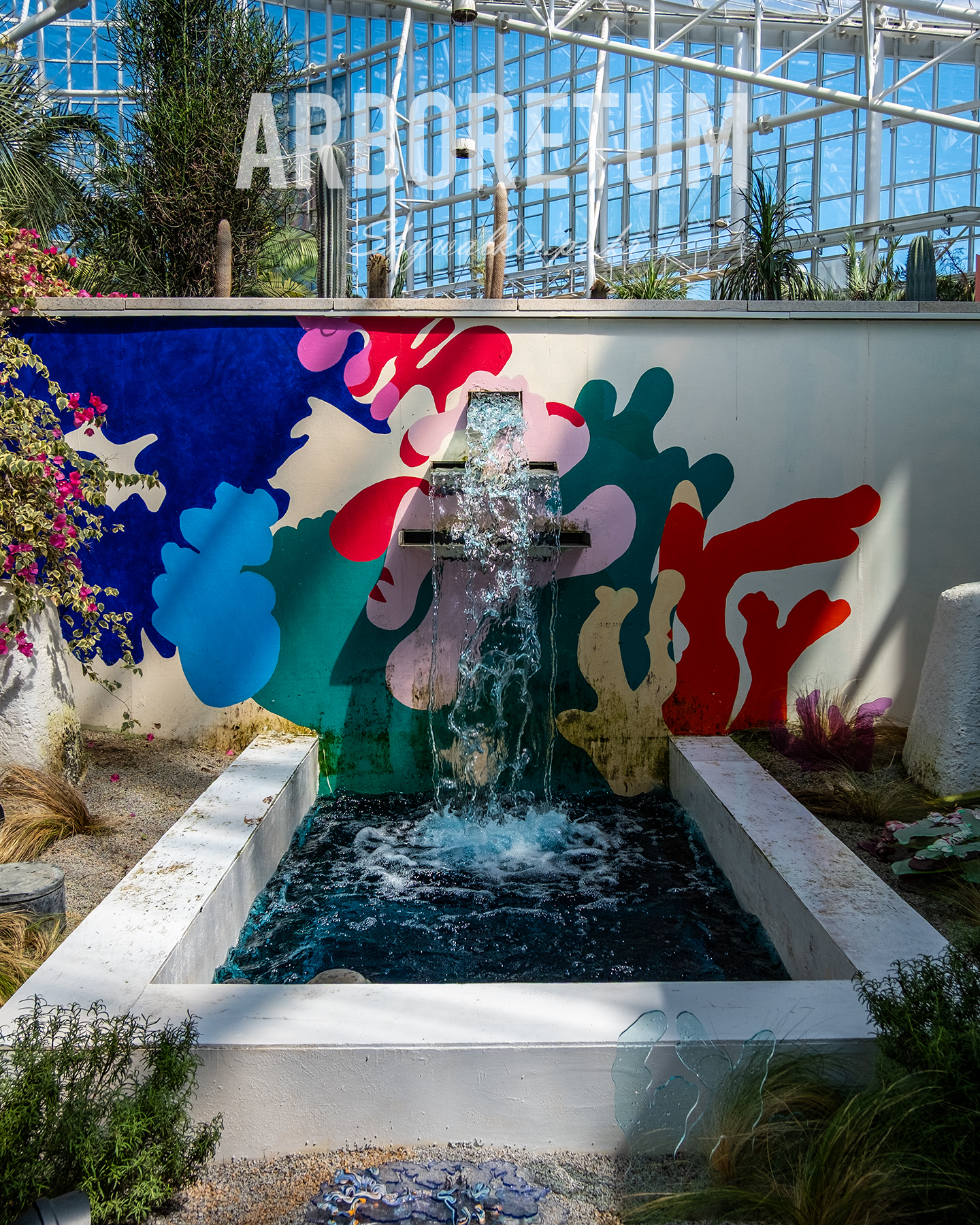 Arboretum - A sunlit conservatory fountain cascades before colorful abstract wall art, framed by desert plants and glass roof beams. - #arboretum #conservatory #fountain #waterfeature #abstractart #desertplants #glasshouse #botanical #sunlight #gardenphotography