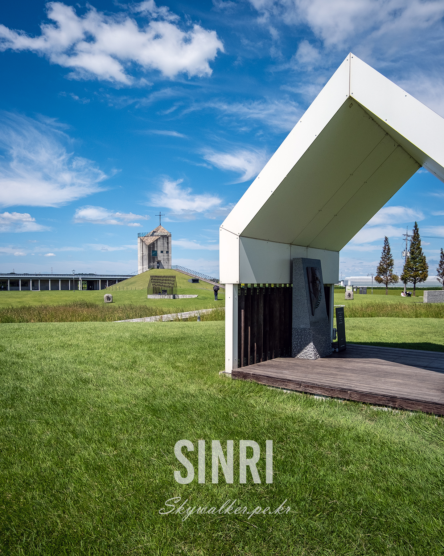 Sinri - A white angular shelter and distant cross-topped tower stand on a green lawn beneath a bright blue sky with scattered clouds. - #landscape #architecture #bluesky #clouds #greenlawn #shelter #tower #minimalism #outdoor #daylight