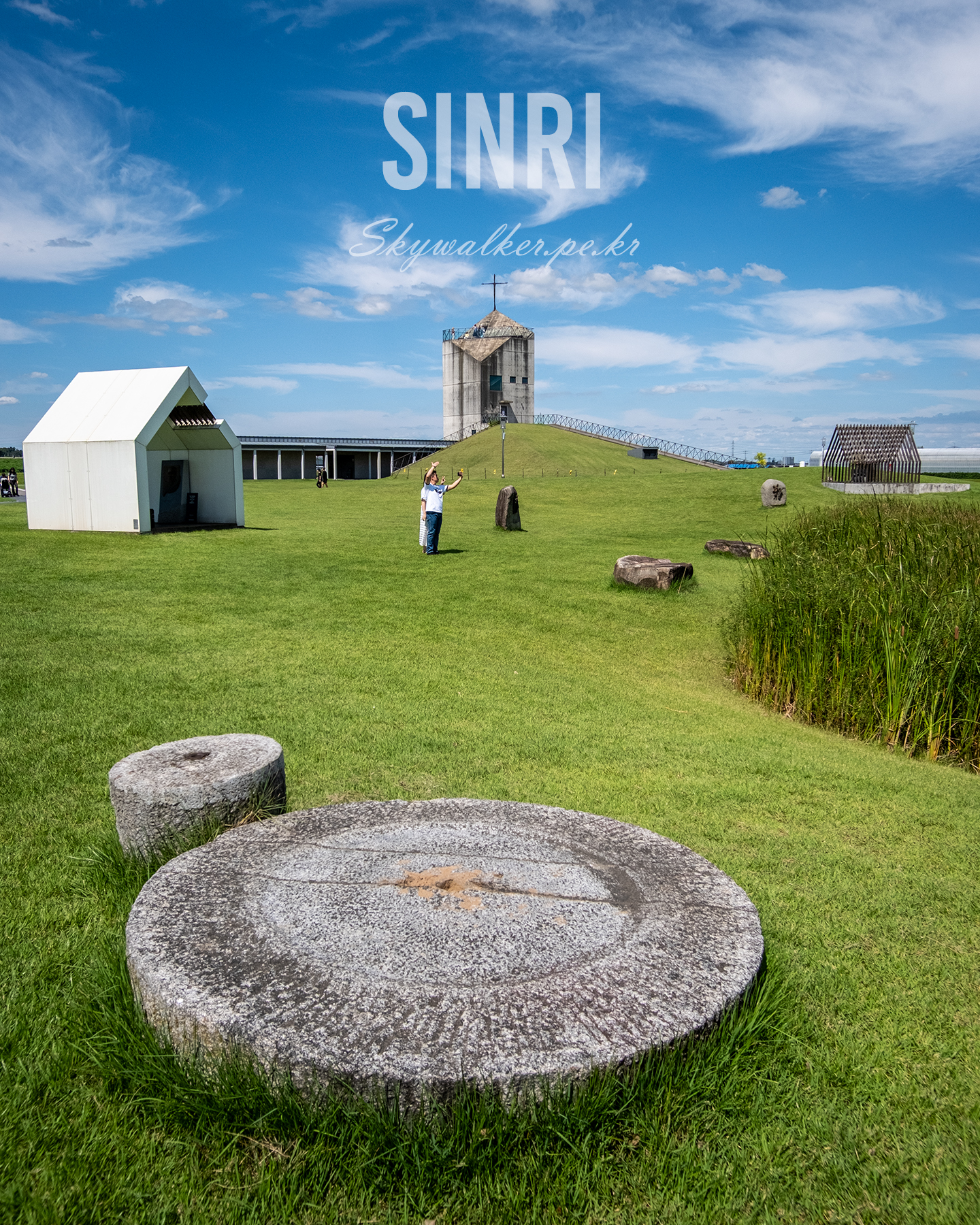 Sinri - A bright grassy park with round stone slabs, small modern structures, and a tower under a vivid blue sky with wispy clouds. - #LandscapePhotography #GreenPark #BlueSky #Cloudscape #Architecture #Grassland #StoneSlabs #SunnyDay #WideAngle #OutdoorScene