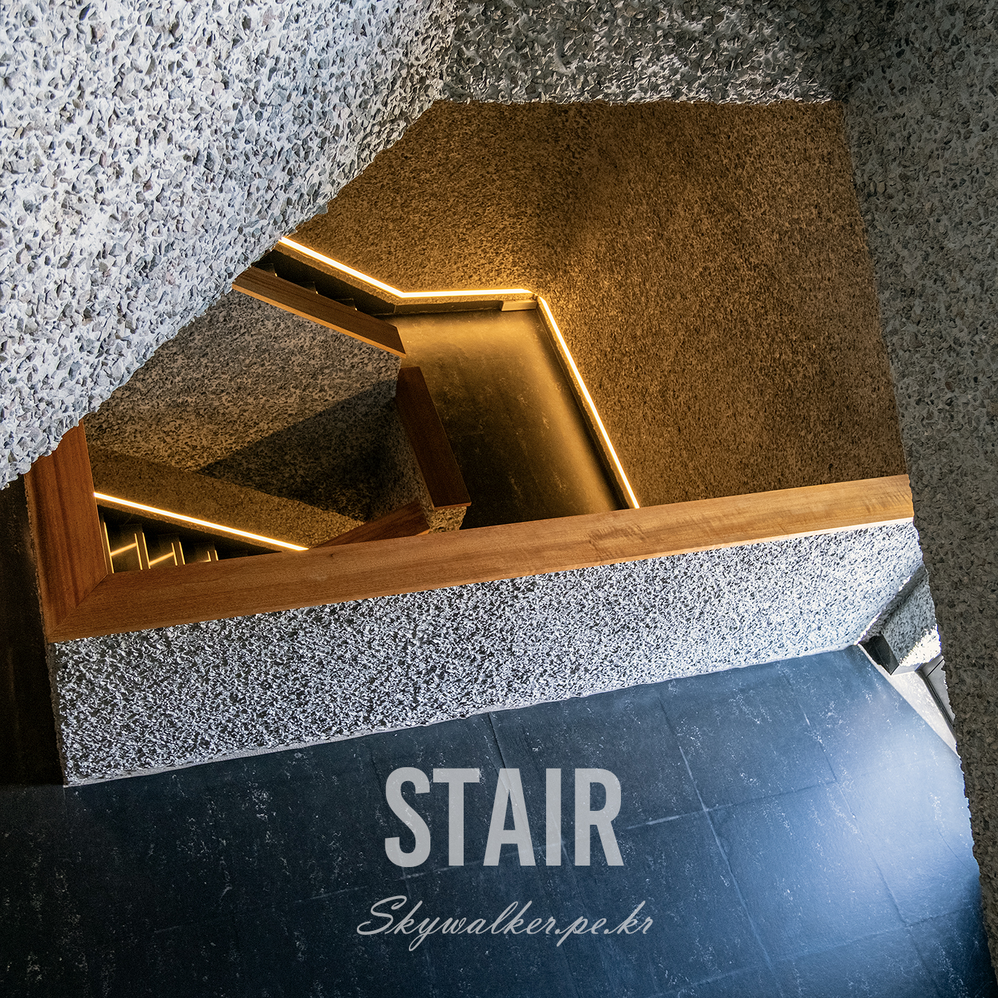 Stair - An angular stairwell descends between rough textured walls, with warm strip lighting and a wooden rail shaping a graphic composition. - #Stair #Stairwell #Architecture #Interior #Concrete #Lighting #ModernDesign #Geometry #WoodRail #UrbanInterior