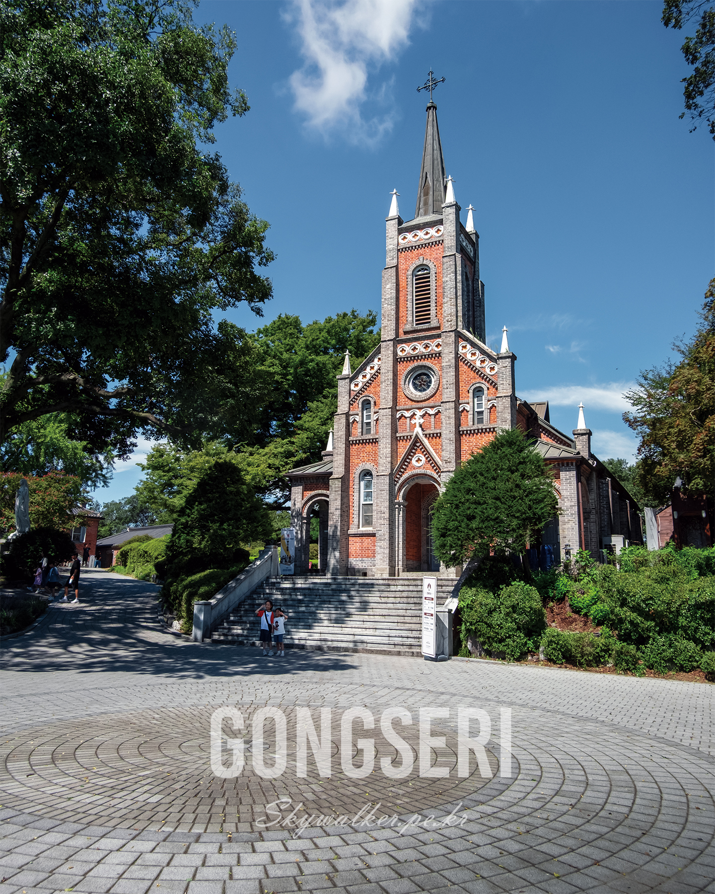 Gongseri - A red brick church with a tall steeple stands among green trees under a clear blue sky in bright daylight. - #church #redbrick #spire #bluesky #trees #architecture #sacredarchitecture #sunlight #landscape #serene