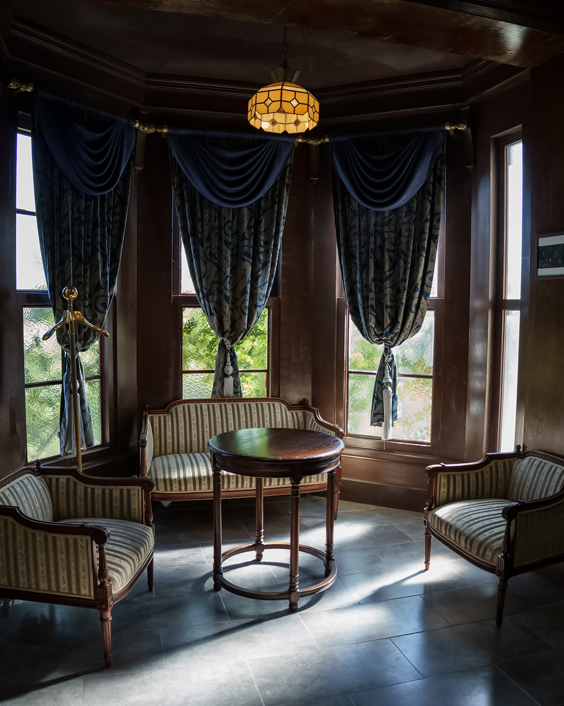 Chairs - An ornate sitting area with upholstered chairs, a round wooden table, tall curtained windows, and warm pendant light in a dark wood room. - #Chairs #VintageInterior #WoodPaneling #RoundTable #BlueDrapes #WindowLight #ClassicDecor #InteriorPhotography #MoodyLight #ElegantRoom