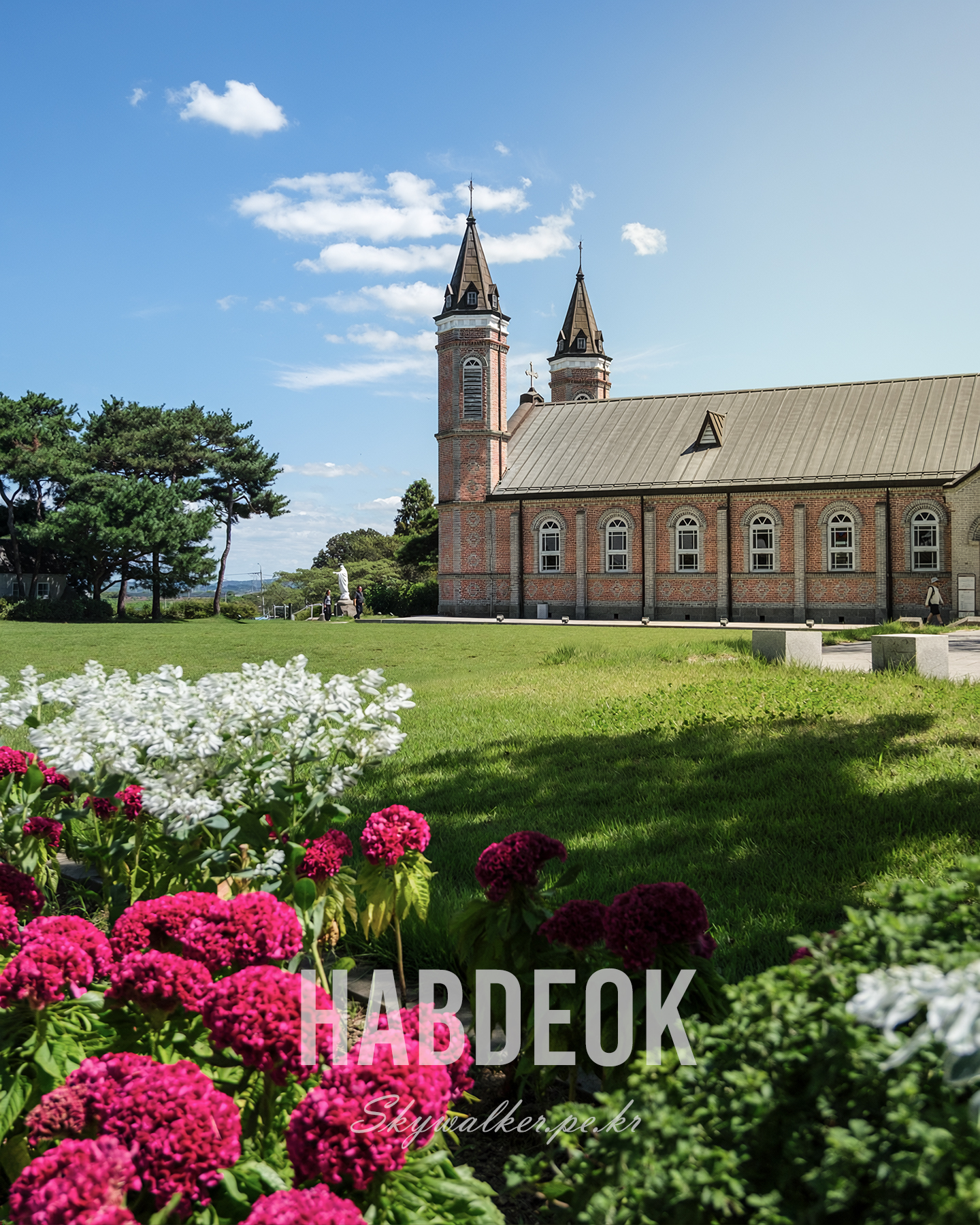 Habdeok - A red brick church with twin spires stands beyond a green lawn and magenta flowers under a clear blue sky. - #Habdeok #BrickChurch #TwinSpires #ChurchArchitecture #FlowerGarden #BlueSky #SunnyDay #GreenLawn #TravelPhotography #ArchitecturalPhotography