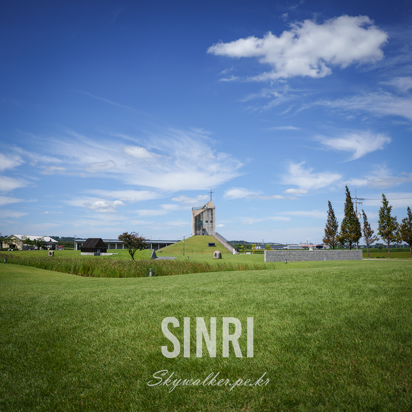 Sinri - A grassy park with a small cross-topped tower sits under a vivid blue sky filled with sweeping white clouds. - #landscape #bluesky #clouds #grassland #chapel #cross #architecture #park #serene #wideangle