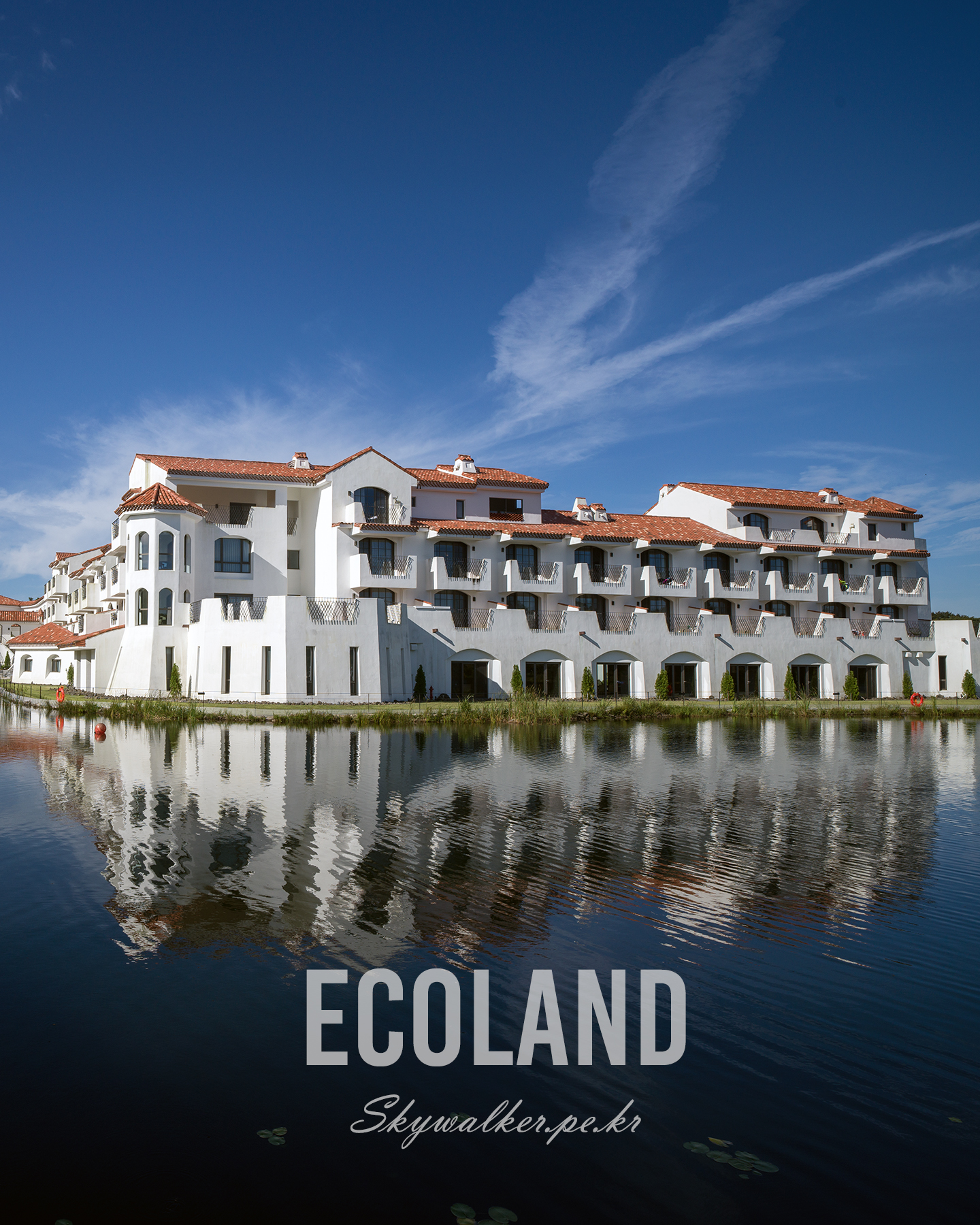 Eco Land - A white red-tiled resort-style building reflects in calm water beneath a deep blue sky with wispy clouds. - #architecture #lakeside #reflection #blueSky #whiteBuilding #redRoof #calmWater #clouds #travelPhotography #serene